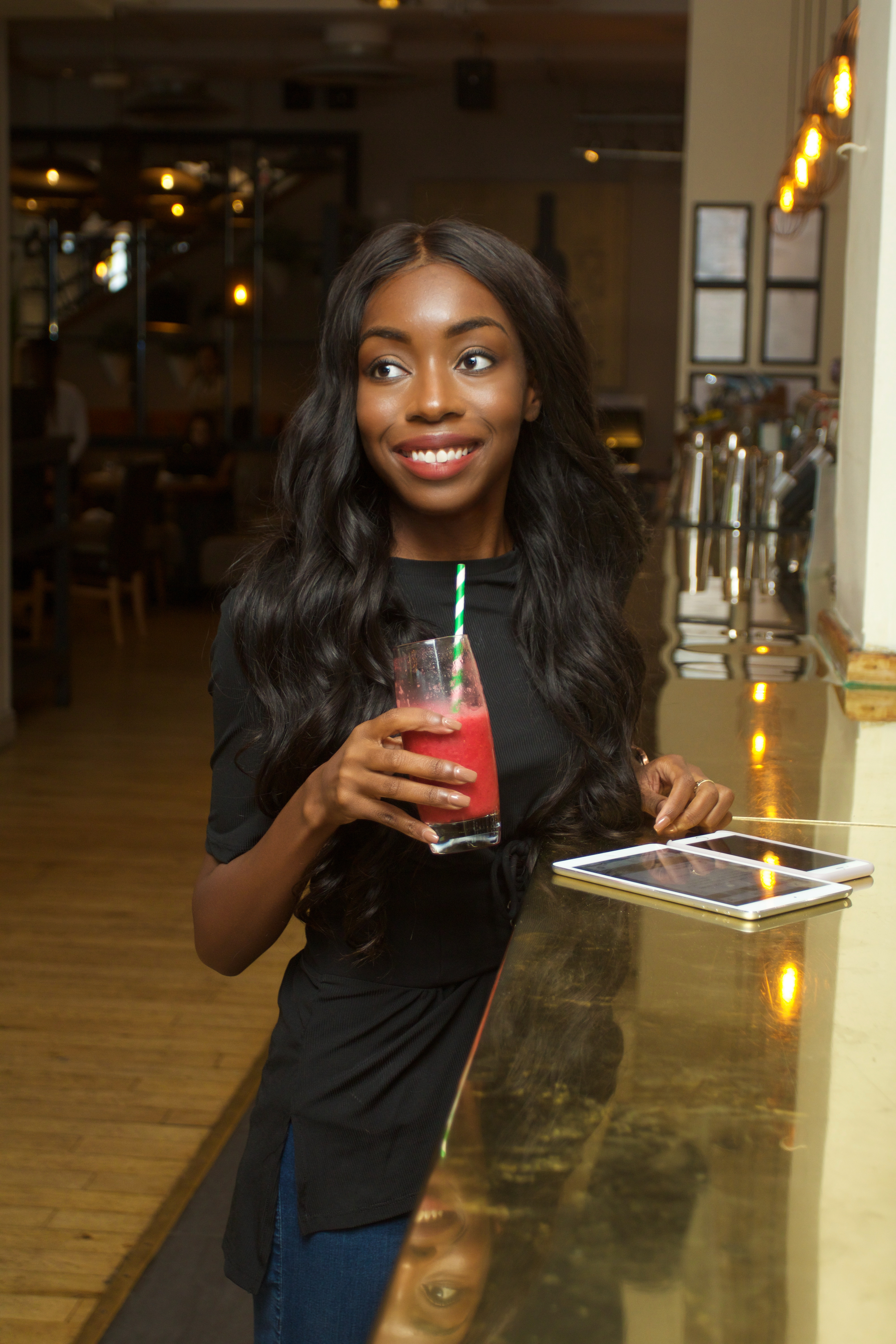 Image of Olivia in a bar, holding a pink drink, with an iPhone and iPad on the bar. Olivia is looking to the side and smiling.